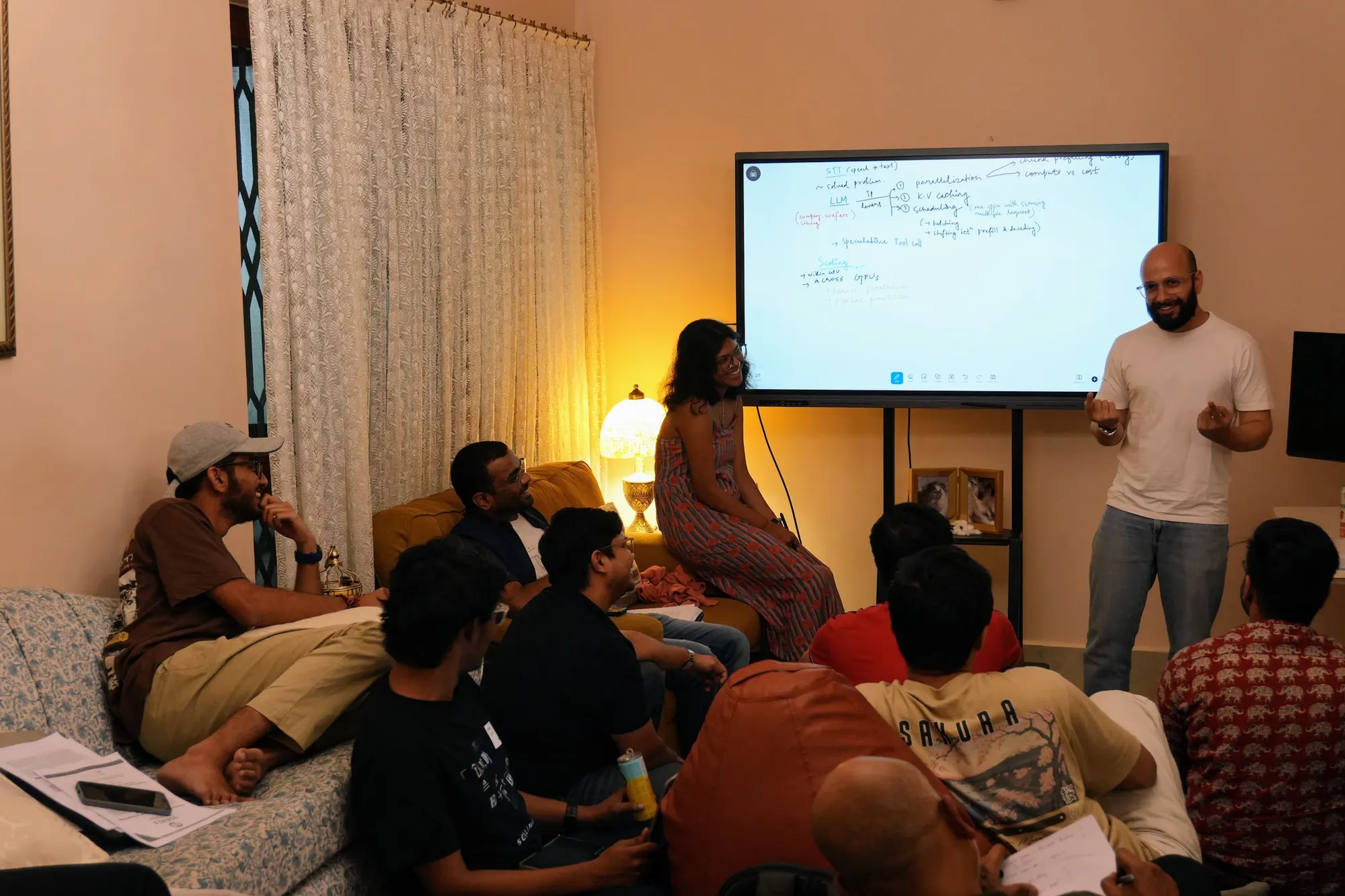The room on sofas and floor cushions while one person talks beside a screen of handwritten notes on LLM levers and scaling.